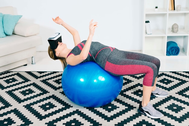 Mujer joven con gafas de realidad virtual que se extiende sobre la bola de pilates tocando las manos en el aire