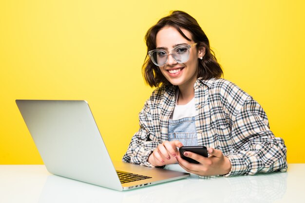 Mujer joven con gafas de sol con las manos usando un teléfono inteligente moderno mientras trabaja en la oficina con computadora aislada