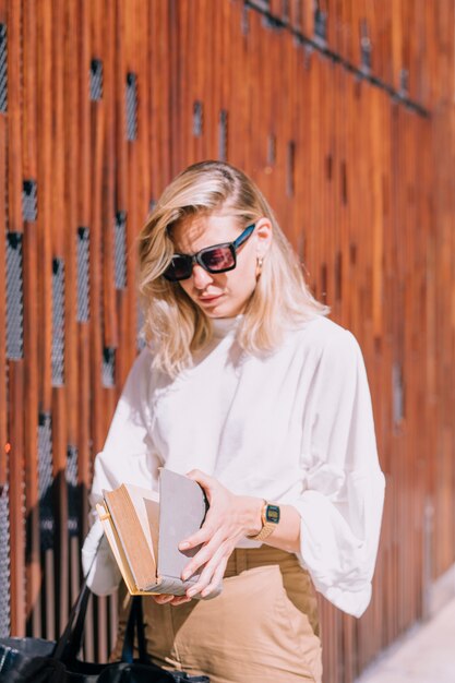 Mujer joven con gafas de sol de pie afuera abriendo el libro