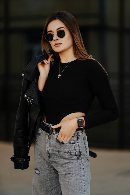 Mujer joven con gafas de sol, sombrero y chaqueta de cuero negra posando al aire libre.