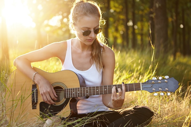 Mujer joven con gafas de sol tocando la guitarra mientras está sentado