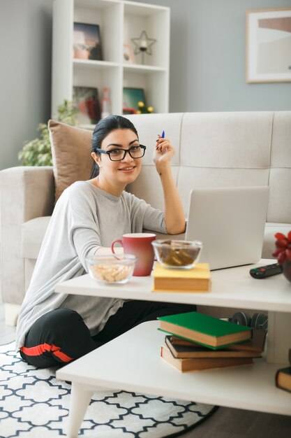 Mujer joven con gafas utiliza portátil sentado en el suelo detrás de la mesa de café en la sala de estar