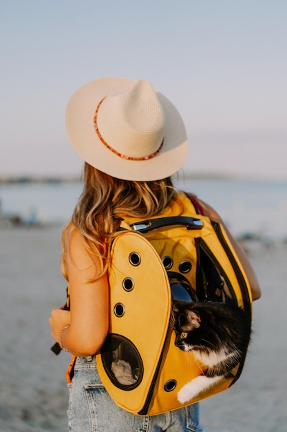 mujer joven con un gato en una mochila a la orilla del mar. Concepto de viaje con una mascota. Gato en la mochila con ojo de buey.