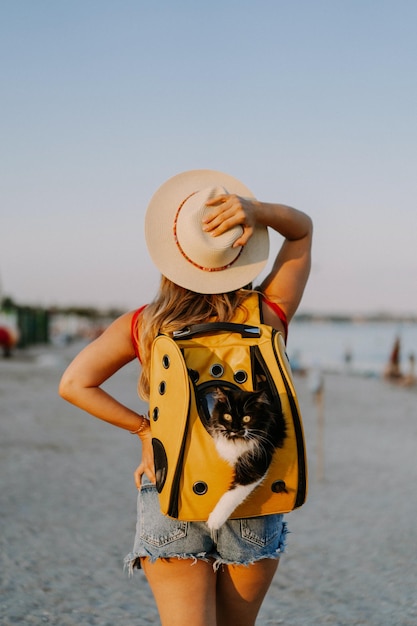 mujer joven con un gato en una mochila a la orilla del mar. Concepto de viaje con una mascota. Gato en la mochila con ojo de buey.