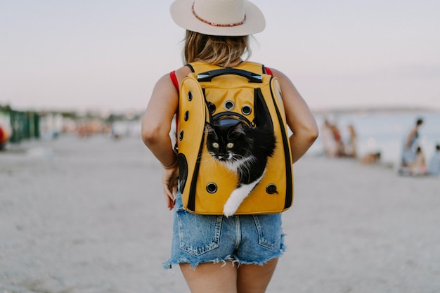 mujer joven con un gato en una mochila a la orilla del mar. Concepto de viaje con una mascota. Gato en la mochila con ojo de buey.