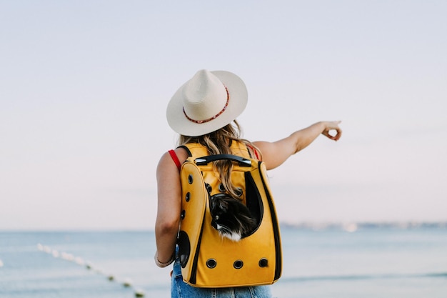 mujer joven con un gato en una mochila a la orilla del mar. Concepto de viaje con una mascota. Gato en la mochila con ojo de buey.