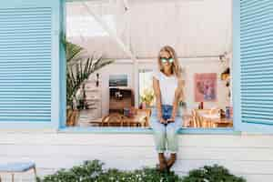 Foto gratuita mujer joven glamorosa en jeans azul sentado en el alféizar de la ventana.