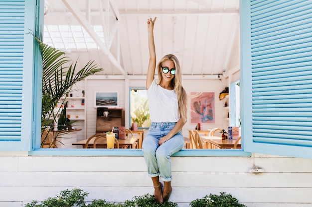 Foto gratuita mujer joven glamorosa en jeans azul sentado en el alféizar de la ventana.