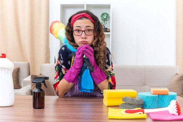 Mujer joven en goma con spray de limpieza y plumero estático mirando guantes confundidos sentado en la mesa con suministros y herramientas de limpieza