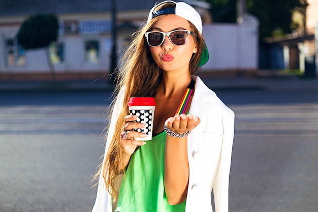 Mujer joven con gorra y sunglasess bebiendo café lanzando un beso en la calle