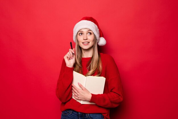 Mujer joven con gorro de Papá Noel sostiene un bloc de notas en sus manos y muerde un bolígrafo revisando la lista de regalos o niños con buen comportamiento aislado en una pared roja.,