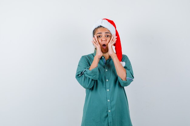 Mujer joven gritando manteniendo las manos cerca de la boca en camisa, gorro de Papá Noel y mirando preocupado, vista frontal.