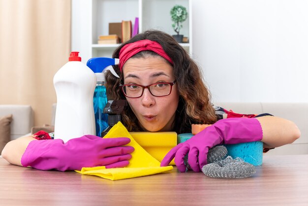 Mujer joven en guantes de goma asombrado y sorprendido sentado en la mesa con artículos de limpieza y herramientas en la sala de luz