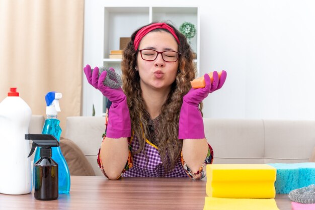 Mujer joven en guantes de goma feliz y positivo que sopla un beso sentado en la mesa con artículos de limpieza y herramientas en la sala de luz