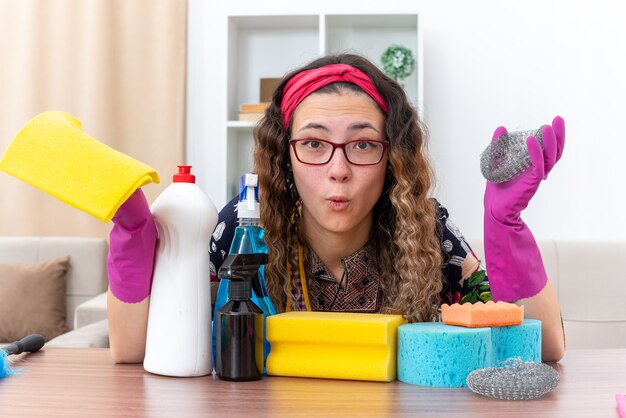 Mujer joven con guantes de goma mirando a la cámara confundida sentada en la mesa con suministros y herramientas de limpieza en la sala de estar ligera