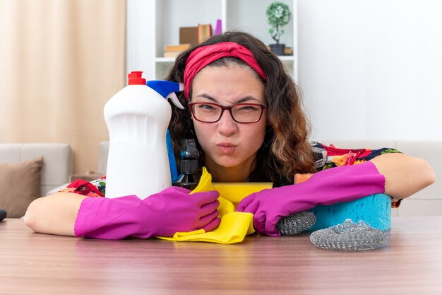 Mujer joven en guantes de goma mirando a la cámara molesto e irritado sentado en la mesa con artículos de limpieza y herramientas en la sala de estar ligera