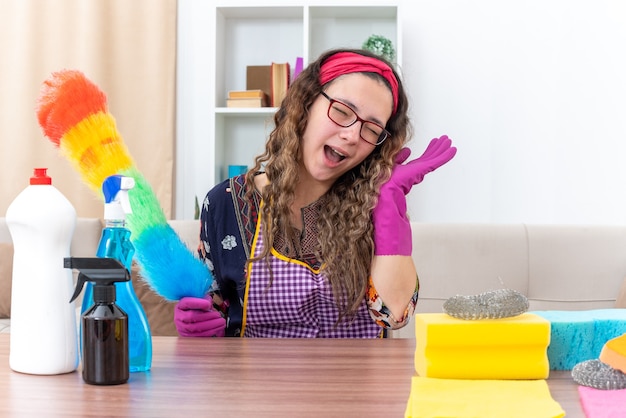 Mujer joven en guantes de goma con plumero estático feliz y alegre sonriendo sentado en la mesa con artículos de limpieza y herramientas en la sala de estar ligera