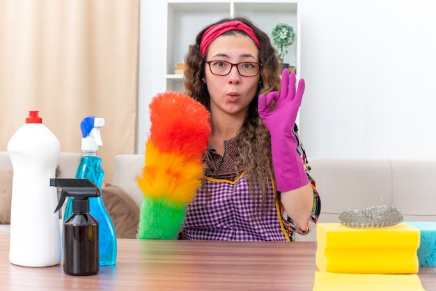 Mujer joven en guantes de goma con plumero estático haciendo signo de ok feliz y positivo sentado en la mesa con suministros y herramientas de limpieza en la sala de estar ligera