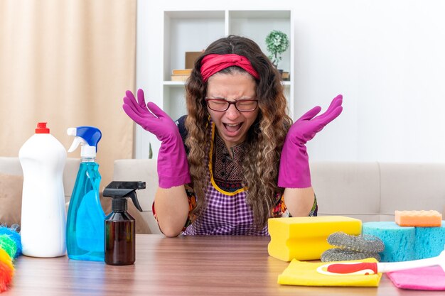 Mujer joven en guantes de goma que parece molesto e irritado gritando y gritando con los brazos levantados sentado en la mesa con artículos de limpieza y herramientas en la sala de luz