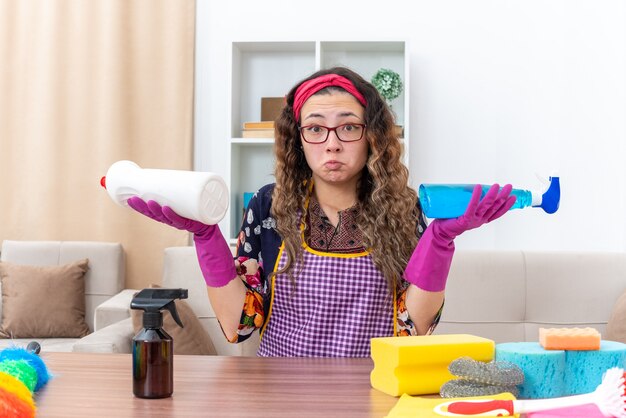 Mujer joven en guantes de goma sentada en la mesa con artículos de limpieza y herramientas confundidas tratando de hacer una elección en la sala de estar ligera