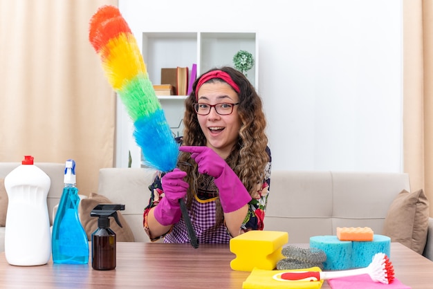 Mujer joven en guantes de goma sosteniendo dusterl estático apuntando con el dedo índice feliz y alegre listo para limpiar sentado en la mesa con artículos de limpieza y herramientas en la sala de estar luminosa