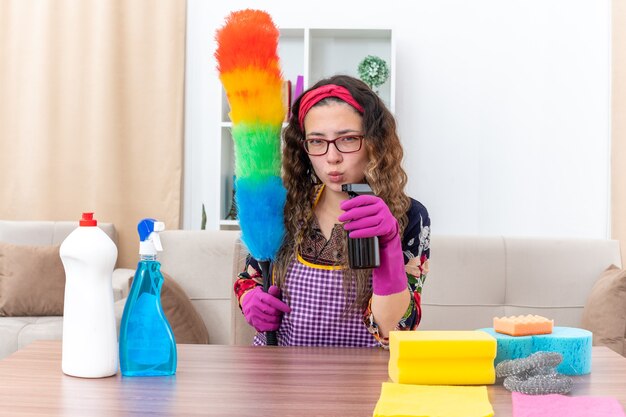 Mujer joven en guantes de goma sosteniendo plumero estático y spray de limpieza mirando confiado sentado en la mesa con suministros y herramientas de limpieza en la sala de estar ligera