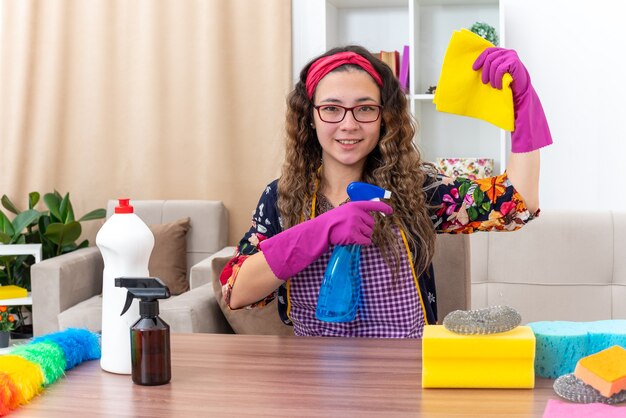 Mujer joven en guantes de goma sosteniendo un trapo y spray de limpieza feliz y positivo sonriendo sentado en la mesa con artículos de limpieza y herramientas en la sala de estar ligera