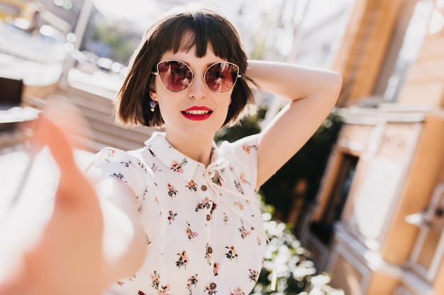 Mujer joven guapa con gafas de sol haciendo selfie en la calle borrosa. Linda chica de pelo corto en blusa de moda pasar tiempo en la ciudad.