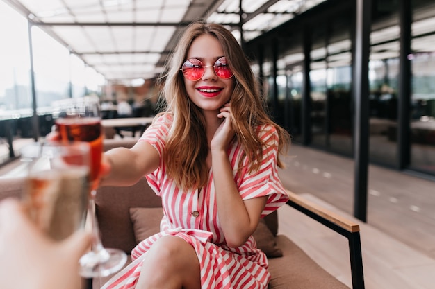 Mujer joven guapa sosteniendo un cóctel y sonriendo en día de verano. Chica rubia extasiada en vasos de color rosa relajante con copa de vino en fin de semana.