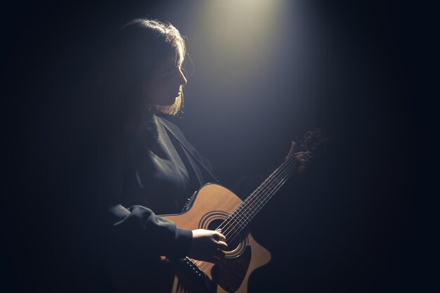 Una mujer joven con una guitarra acústica en la oscuridad bajo un rayo de luz.