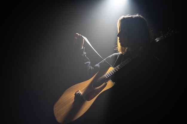 Una mujer joven con una guitarra acústica en la oscuridad bajo un rayo de luz.