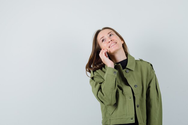 Mujer joven hablando con alguien por teléfono móvil en chaqueta verde y mirando feliz. vista frontal. espacio para texto