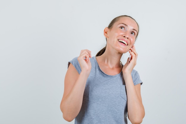 Mujer joven hablando por teléfono celular en camiseta gris y mirando alegre, vista frontal.