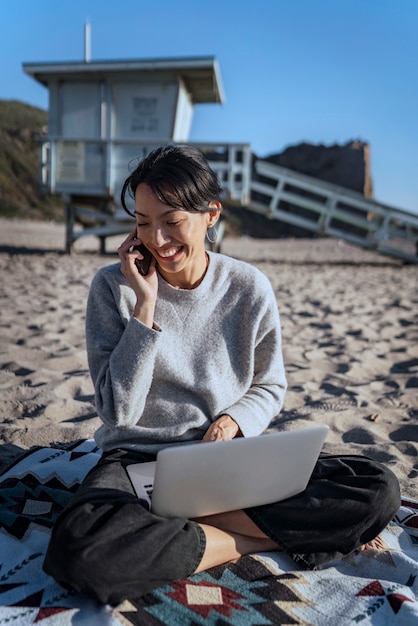 Mujer joven hablando por teléfono inteligente mientras usa su computadora portátil en la playa
