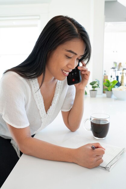 Mujer joven hablando por teléfono inteligente y tomando una taza de café mientras toma notas