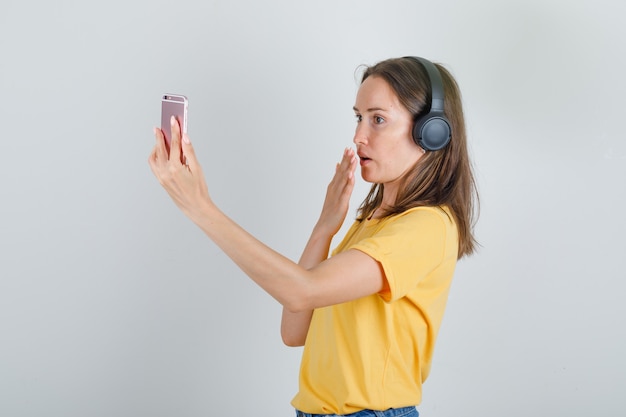 Mujer joven hablando por teléfono inteligente a través de videollamada en camiseta amarilla