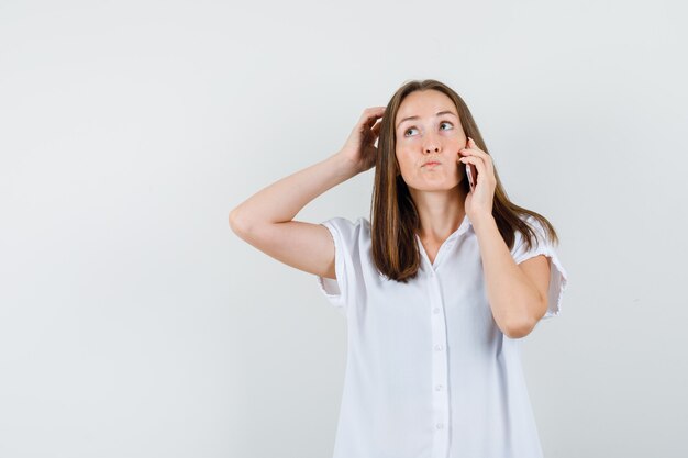 Mujer joven hablando por teléfono mientras se rasca la cabeza con una blusa blanca y mira pensativa.