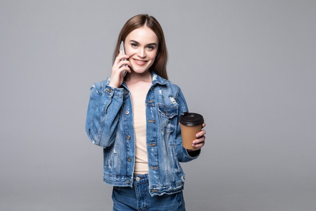 Mujer joven hablando por teléfono móvil bebiendo café posando aislado sobre pared gris