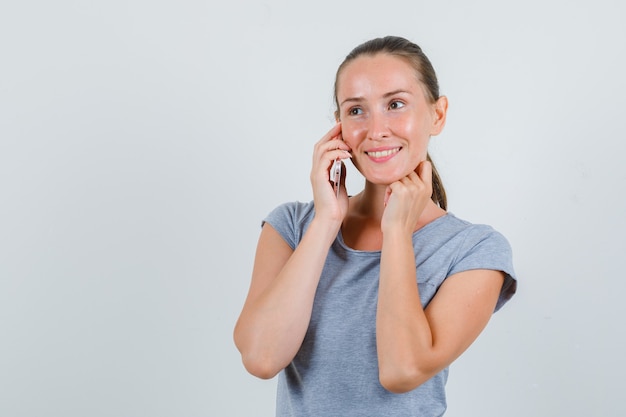 Mujer joven hablando por teléfono móvil en camiseta gris y mirando alegre. vista frontal.