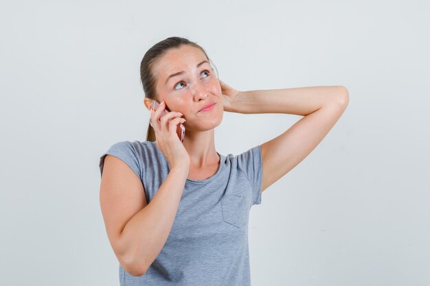 Mujer joven hablando por teléfono móvil en camiseta gris y mirando vacilante, vista frontal.