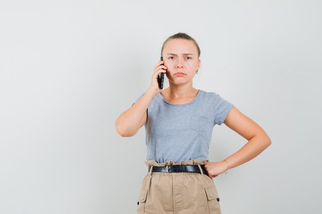 Mujer joven hablando por teléfono móvil en camiseta, pantalón y mirando vacilante. vista frontal.