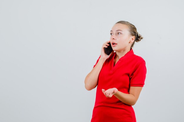 Mujer joven hablando por teléfono móvil en camiseta roja y mirando confundido