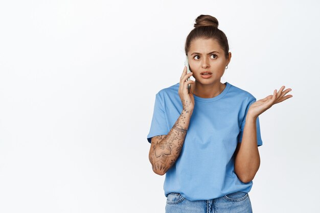 Mujer joven hablando por teléfono móvil con cara de confusión, de pie en camiseta azul sobre blanco