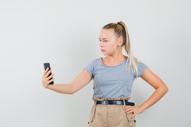 Mujer joven hablando por video chat en camiseta, pantalón y mirando serio. vista frontal.