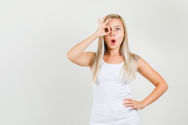 Mujer joven haciendo bien firmar con la mano en el ojo en camiseta blanca y mirando positivo