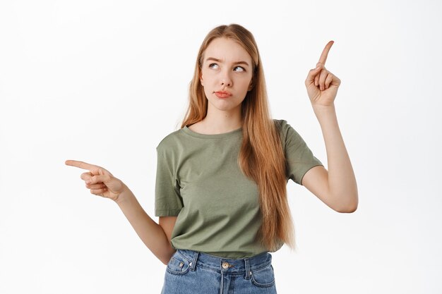 Mujer joven haciendo elección entre dos lados, apuntando hacia los lados y mirando pensativa, reflexionando sobre su decisión, necesita consejo sobre qué elegir, de pie sobre una pared blanca