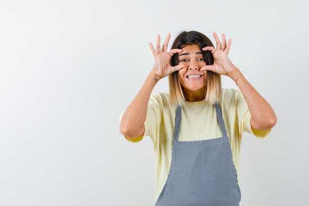 Mujer joven haciendo un gesto de mano de caja en sus ojos y sonriendo sobre fondo blanco.