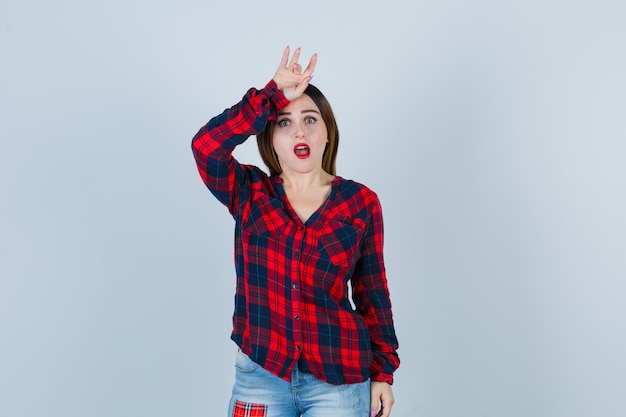 Mujer joven haciendo un gesto con la mano por encima de la cabeza, manteniendo la boca abierta en camisa a cuadros, jeans y mirando sorprendido, vista frontal.