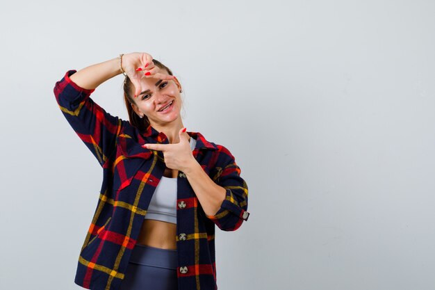 Foto gratuita mujer joven haciendo gesto de marco en la parte superior de la cosecha, camisa a cuadros y mirando feliz. vista frontal.