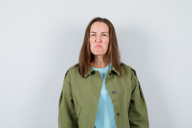 Mujer joven haciendo muecas en chaqueta verde y mirando gruñón, vista frontal.